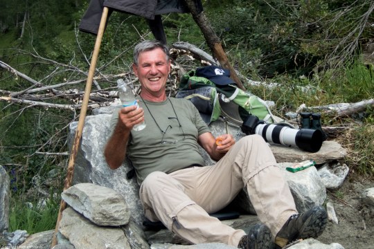 LBV Ehrenamtlicher Richard Straub sitzt auf einem Felsvorsprung und lacht. Er hält eine Wasserflasche hoch und neben ihm liegt eine Kamera mit langem Teleobjektiv | © Richard Straub