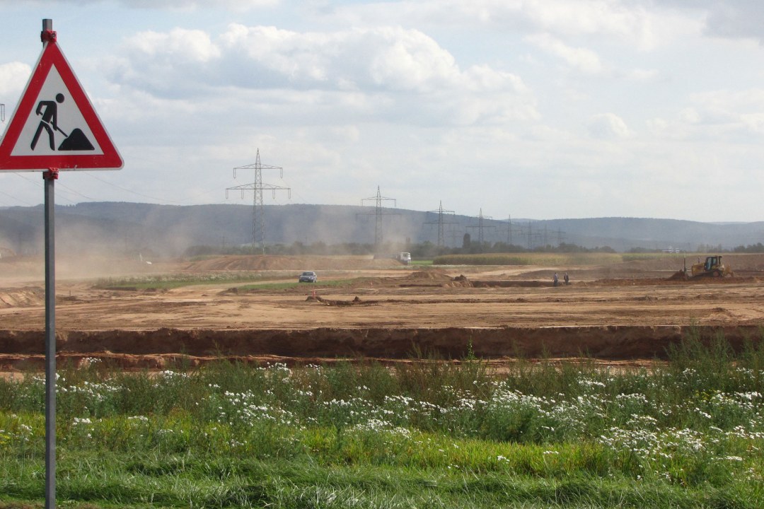 Grüne Wiese im Vordergrund, dahinter eine planierte Fläche mit Autos und Traktoren. Links steht ein Baustellenschild | © Thomas Staab