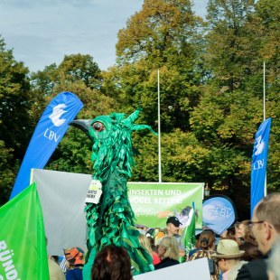 Stelzentänzer als grüner Vogel verkleidet auf der Demo Mia ham's satt zwischen Menschen und Bannern | © LBV