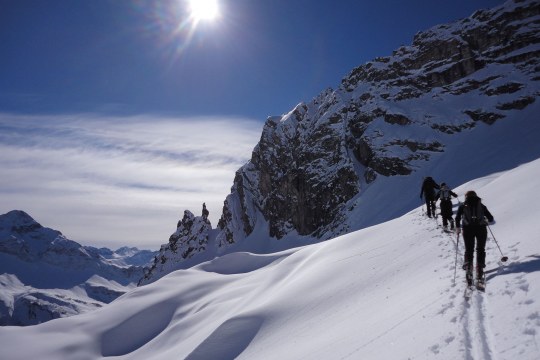 Drei Menschen nacheinander auf einer Skitour in Oberstdorf. Es liegt viel Schnee in den zu sehenden Bergen, die Sonne scheint am blauen Himmel | © Henning Werth